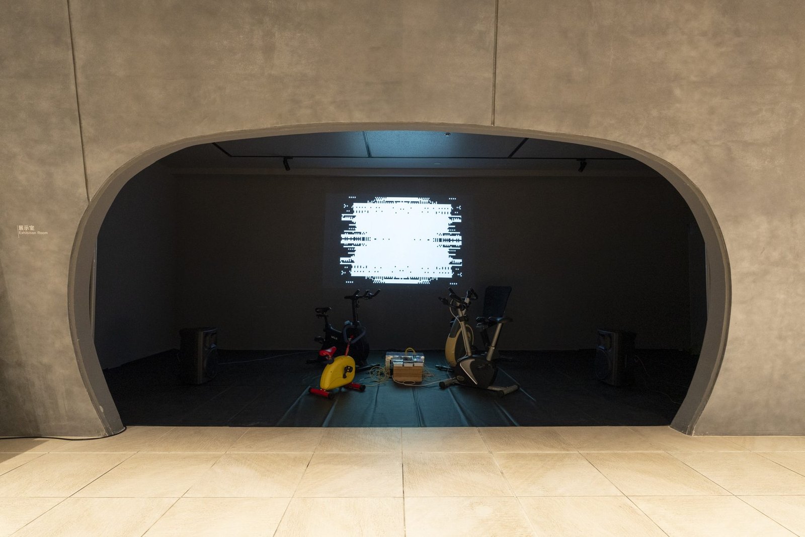 Velophone at NAHArT: four exercise bikes, one of them a small yellow children’s bike, gathered in a dark room before a projected black-and-white video-synth image, seen through an arched doorway.